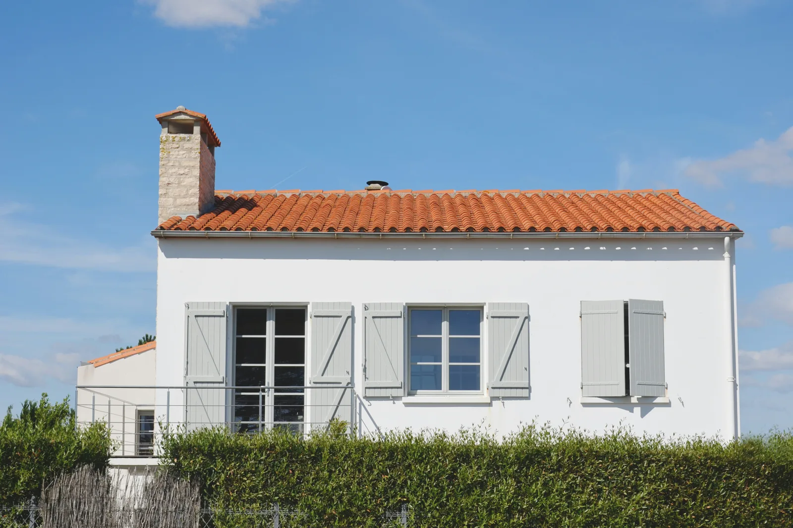 Maison blanche aux volets gris et au toit de tuiles, derrière une haie, sous un ciel bleu